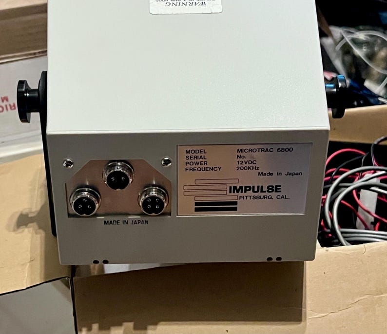 May include: A light gray electronic device with a metal casing, featuring three circular ports on the front panel. A label displays the text: MODEL MICROTRAC 6800, POWER 12VDC, FREQUENCY 200KHz, IMPULSE PITTSBURG, CAL., and MADE IN JAPAN.