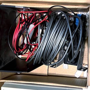 May include: A cardboard box containing a collection of electronic components, including a large coil of grey cable, several red and black insulated wires, a black plastic housing, and metal mounting brackets for marine transducer equipment.