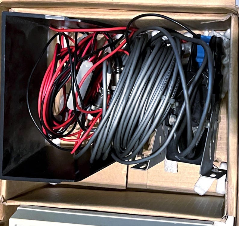 May include: A cardboard box containing a collection of electronic components, including a large coil of grey cable, several red and black insulated wires, a black plastic housing, and metal mounting brackets for marine transducer equipment.