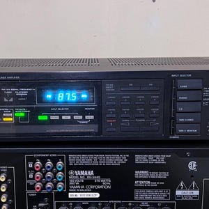 Può includere: Amplificatore sintonizzatore sintetizzato al quarzo Onkyo nero con display digitale che mostra "87.5". L'unità ha vari pulsanti e manopole per la selezione dell'ingresso e il controllo del volume. Sotto, un ricevitore Yamaha nero.