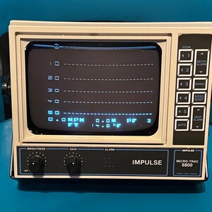 May include: An off-white Impulse Micro-Trac 6800 video fish finder with a black control panel. The screen displays a digital depth grid with readings of 0.0 MPH, 0.0M, and 14.2 F. It features knobs for brightness and gain, plus side buttons.