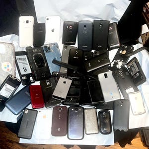 May include: A collection of various smartphones, in different colors and conditions, including white, black, and silver models. Some phones have cracked screens or missing components, while others appear intact. The phones are arranged on a white surface.