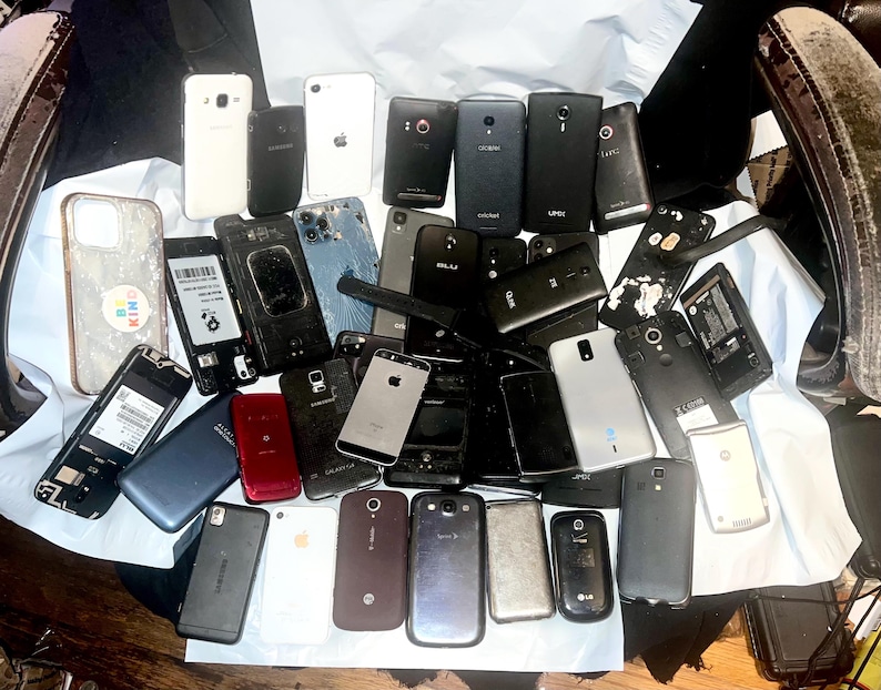 May include: A collection of various smartphones, in different colors and conditions, including white, black, and silver models. Some phones have cracked screens or missing components, while others appear intact. The phones are arranged on a white surface.