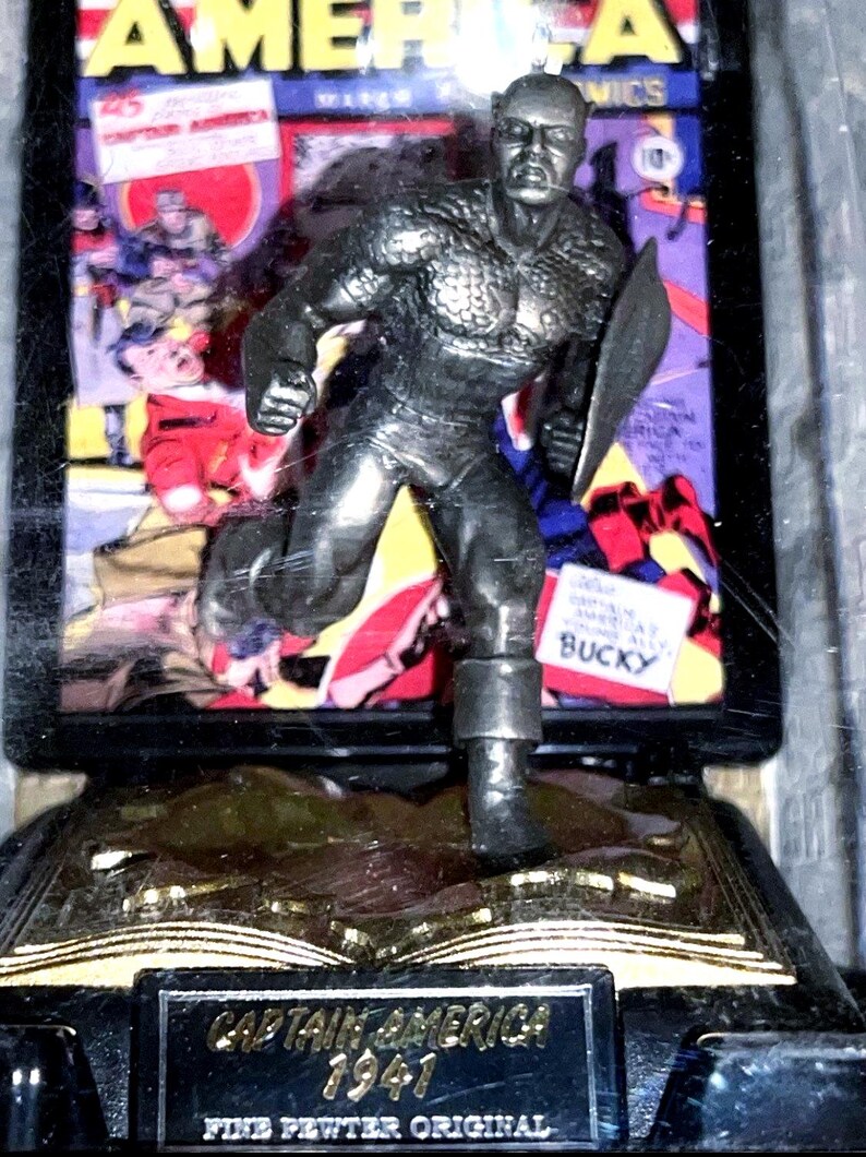 K&ouml;nnte beinhalten: Eine Captain America Figur aus feinem Zinn. Die Figur befindet sich in einer Laufpose und h&auml;lt einen Schild. Auf der Basis stehen die Worte "Captain America 1941" und "Fine Pewter Original". Im Hintergrund ist ein Comic-Cover zu sehen.
