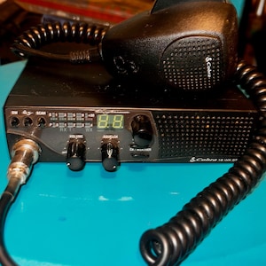 Peut inclure: Un émetteur-récepteur CB Cobra 18 WX ST noir avec un écran numérique affichant 8.8. La radio possède un microphone avec un cordon enroulé et plusieurs boutons, notamment le volume, le squelch et le CB/météo.