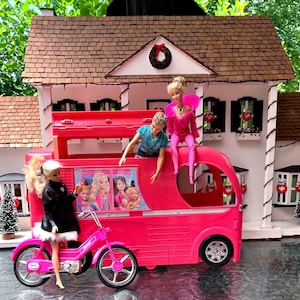 Op de afbeelding: Een felroze speelgoedcamper met een afbeelding van vier poppen op de zijkant, met één pop zittend op het dak, één pop leunend uit de bovenste opening en één pop staand naast een bijpassende roze fiets voor een poppenhuis.