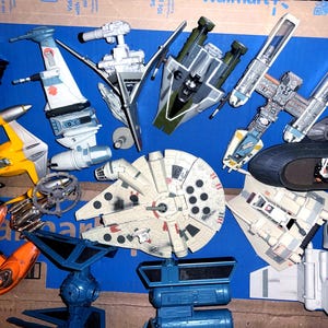 Könnte beinhalten: Eine Sammlung von Star Wars Spielzeugraumschiffen und -fahrzeugen in verschiedenen Farben. Das Sortiment umfasst den Millennium Falken, X-Wings, Y-Wings und andere ikonische Raumschiffe aus dem Star Wars-Franchise. Die Spielzeuge sind auf einer blauen Oberfläche angeordnet.