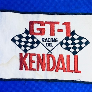 May include: A white patch with a black border featuring the text "GT-1 RACING OIL KENDALL" in red. Two crossed checkered flags are also embroidered in black and white. The patch is set against a blue background.