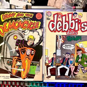 Op de afbeelding: Twee vintage stripboeken. De eerste, "Danny and the Demoncycle", toont een jonge man op een oranje motorfiets. De tweede, "Debbi's Dates", heeft een retro-ontwerp met het woord "THUMP!" en de tekst "Daddy rigged that up buddy!"