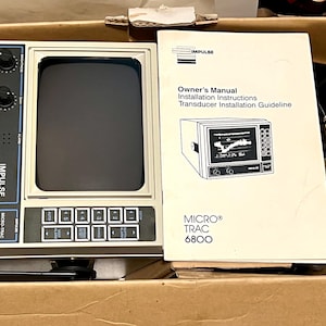 May include: An Impulse Micro-Trac 6800 marine fish finder unit with a beige casing, black screen, and control panel featuring knobs and buttons, accompanied by an owner's manual and installation instructions booklet inside a cardboard box.