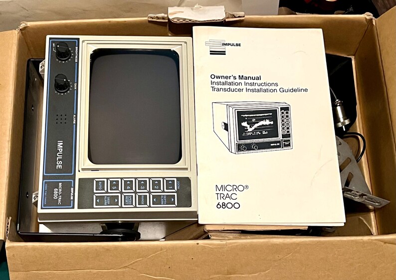 May include: An Impulse Micro-Trac 6800 marine fish finder unit with a beige casing, black screen, and control panel featuring knobs and buttons, accompanied by an owner's manual and installation instructions booklet inside a cardboard box.