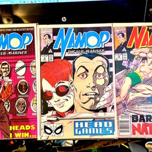 May include: Three comic book covers featuring the superhero Namor, the Sub-Mariner. The covers are red, blue, and yellow with black text. The first cover features Namor in a red and black suit, the second cover features Namor with a woman with red hair and a black eye mask, and the third cover features Namor in a green and black swimsuit.