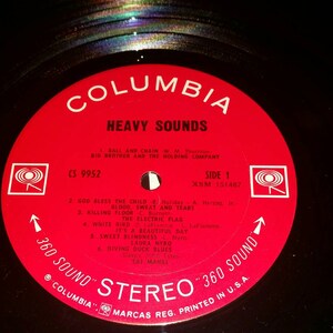 May include: A black vinyl record with a circular red Columbia label. The label features the text HEAVY SOUNDS, track listings, and the 360 SOUND STEREO logo. The record has a central spindle hole and shows a rainbow-colored light reflection.