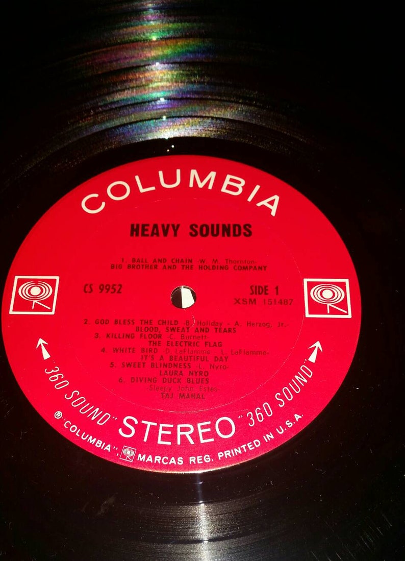 May include: A black vinyl record with a circular red Columbia label. The label features the text HEAVY SOUNDS, track listings, and the 360 SOUND STEREO logo. The record has a central spindle hole and shows a rainbow-colored light reflection.