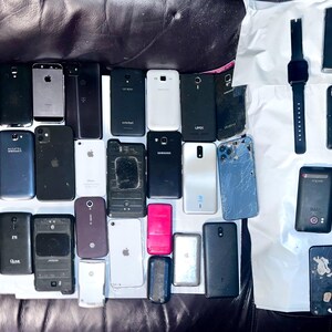 May include: A collection of various smartphones and a smartwatch, in different colors and designs, arranged on a dark surface. The phones vary in size and style, with some showing signs of wear.