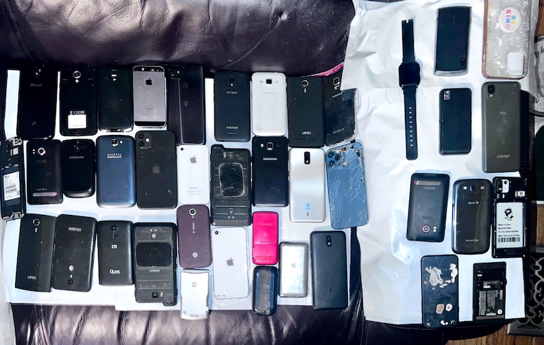 May include: A collection of various smartphones and a smartwatch, in different colors and designs, arranged on a dark surface. The phones vary in size and style, with some showing signs of wear.