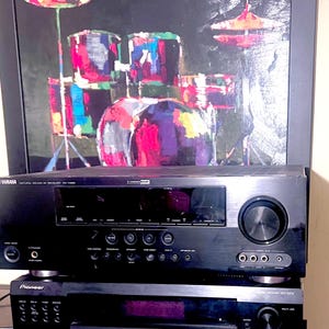 以下が含まれることがあります： 黒いヤマハレシーバー。複数のボタンと大きなダイヤルがあり、黒いパイオニアレシーバーの上に積み重ねられています。ドラムセットのカラフルな絵が背景に飾られています。