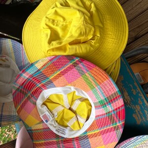 May include: A selection of sun hats in different designs. The top hat is a vibrant yellow, featuring a broad brim. Beneath it, a hat displays a colourful plaid pattern. The interior of the plaid hat is yellow and white, with a label that says "Town Talk".