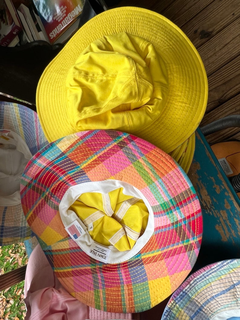 May include: A selection of sun hats in different designs. The top hat is a vibrant yellow, featuring a broad brim. Beneath it, a hat displays a colourful plaid pattern. The interior of the plaid hat is yellow and white, with a label that says "Town Talk".