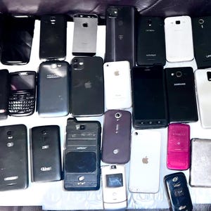 May include: A collection of various smartphones, in black, white, and silver, with different designs and brands. Some phones have cracked screens. The phones are arranged on a dark surface.