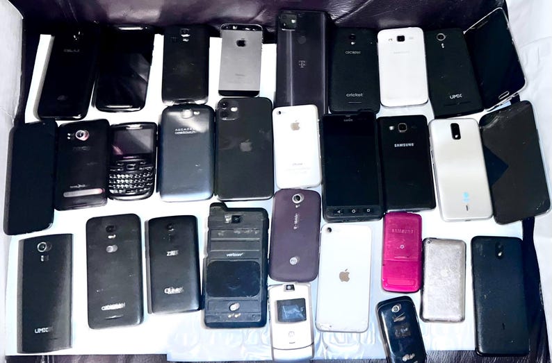 May include: A collection of various smartphones, in black, white, and silver, with different designs and brands. Some phones have cracked screens. The phones are arranged on a dark surface.