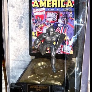K&ouml;nnte beinhalten: Feine Zinnfigur von Captain America in einer transparenten Verpackung mit Comic-Cover-Hintergrund. Die Verpackung tr&auml;gt die Aufschrift "Comic Book Champions", "Marvel Comics" und "Golden Age".