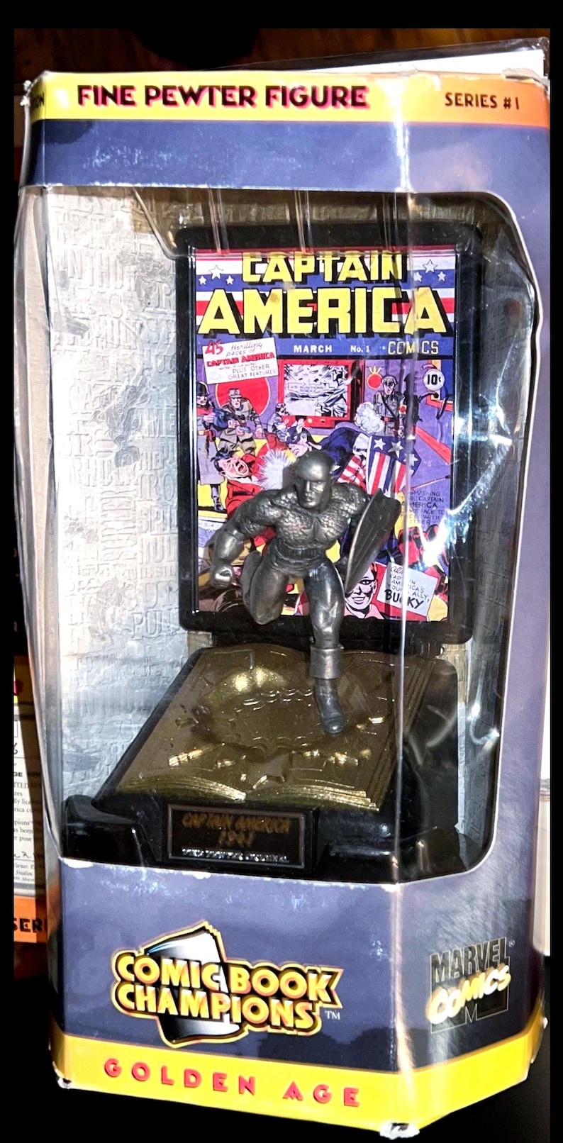 K&ouml;nnte beinhalten: Feine Zinnfigur von Captain America in einer transparenten Verpackung mit Comic-Cover-Hintergrund. Die Verpackung tr&auml;gt die Aufschrift "Comic Book Champions", "Marvel Comics" und "Golden Age".