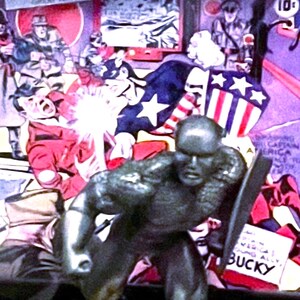 K&ouml;nnte beinhalten: Eine metallische Captain America-Figur in dynamischer Pose, die einen Schild h&auml;lt. Der Hintergrund zeigt ein Vintage-Comic-Cover mit dem Titel "Captain America Comics" und dem Wort "March". Das Comic-Cover hat ein patriotisches Thema.