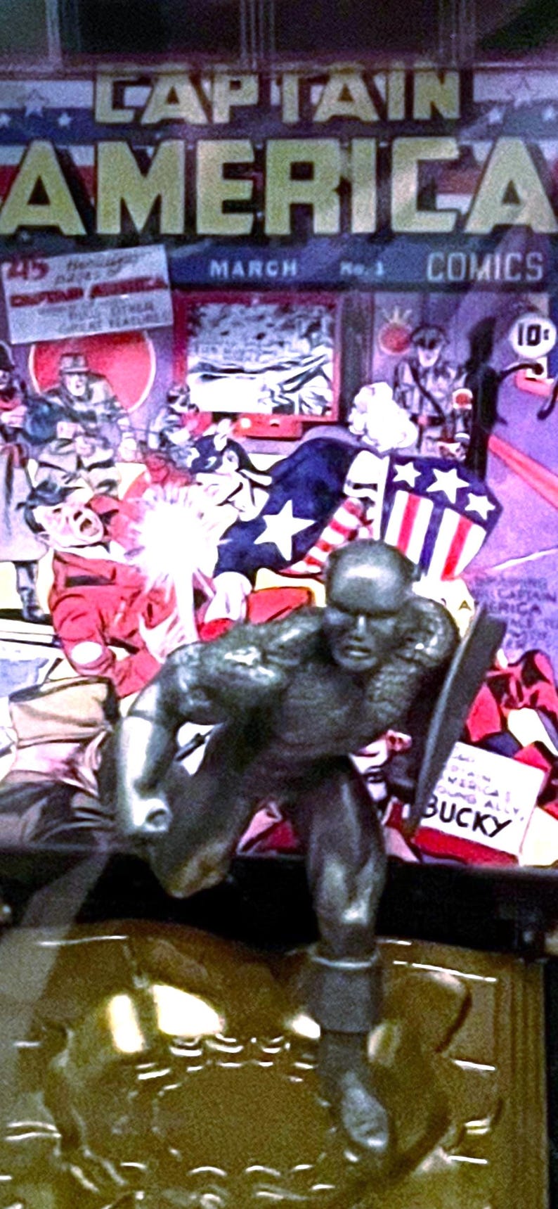 K&ouml;nnte beinhalten: Eine metallische Captain America-Figur in dynamischer Pose, die einen Schild h&auml;lt. Der Hintergrund zeigt ein Vintage-Comic-Cover mit dem Titel "Captain America Comics" und dem Wort "March". Das Comic-Cover hat ein patriotisches Thema.