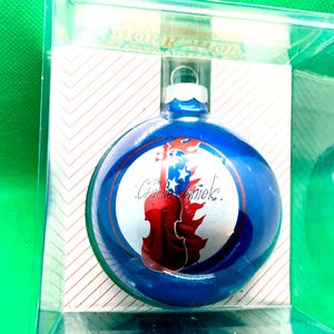 May include: A round blue glass Christmas ornament encased in a clear plastic box. The ornament features a central white circular graphic depicting a red violin silhouette against a blue background with white stars and the text The Charlie Daniels.