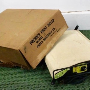 Op de afbeelding: Een vintage Premier Print Dryer Model TC van Photo Materials Co. met de originele bruine kartonnen doos. Het apparaat heeft een crèmekleurig canvas oppervlak, een zwart metalen frame en een geel bedieningspaneel met een zwarte knop.