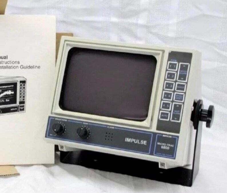 May include: An Impulse Micro-Trac 6800 marine electronic display unit with a gray casing, a dark screen, and a black control panel featuring buttons and knobs. The device includes a mounting bracket with side adjustment knobs and an instruction manual.