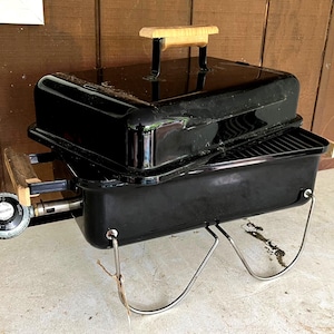 Könnte beinhalten: Ein schwarzer tragbarer Holzkohlegrill mit Deckel und Holzgriff. Der Grill hat einen verchromten Ständer und einen Gasregler. Der Grill ist für das Kochen und Grillen im Freien konzipiert.