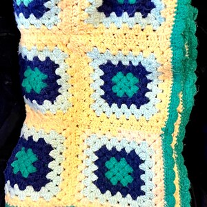May include: Handmade crochet blanket featuring a granny square design. The blanket has a yellow base with squares in shades of blue, teal, and white. A teal scalloped edge frames the blanket. This cozy blanket is perfect for a bed or sofa.