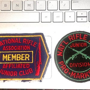May include: Two vintage patches. One is a navy blue hexagon with red text that reads "NATIONAL RIFLE ASSOCIATION MEMBER AFFILIATED JUNIOR CLUB" and two red rifles. The other is a black circle with green text that reads "NAT'L RIFLE ASSN JUNIOR DIVISION PRO-MARKSMAN".