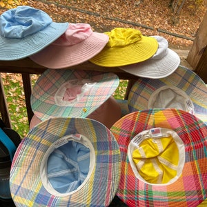 May include: A selection of sun hats in shades of blue, pink, yellow, and white. Some hats display a pastel plaid design. The hats have broad brims and are made from a light material, ideal for shielding from the sun.