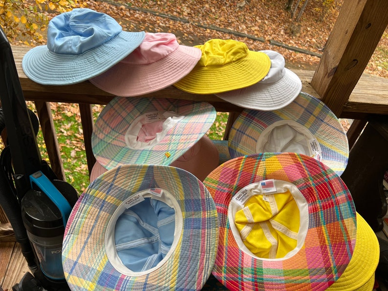 May include: A selection of sun hats in shades of blue, pink, yellow, and white. Some hats display a pastel plaid design. The hats have broad brims and are made from a light material, ideal for shielding from the sun.