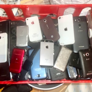 May include: A red plastic crate filled with various smartphones and a smartwatch. The phones are in different colors, including white, black, silver, and red. Some phones have the Apple logo.