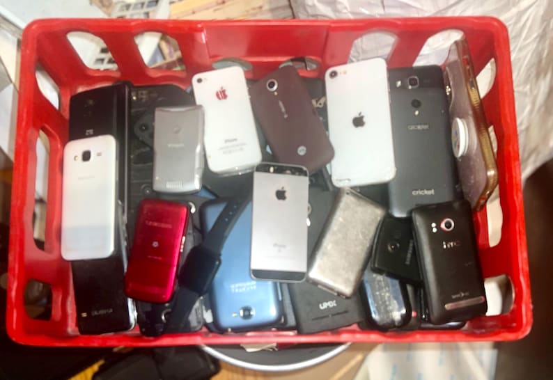 May include: A red plastic crate filled with various smartphones and a smartwatch. The phones are in different colors, including white, black, silver, and red. Some phones have the Apple logo.