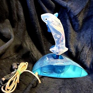 Könnte beinhalten: Ein durchsichtiger Joystick in Delfinform mit blauem Sockel. Der Joystick hat einen blauen Knopf und ein USB-Kabel. Der Sockel ist transparent blau. Auf schwarzem Hintergrund.