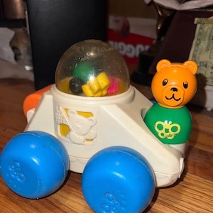 May include: A white plastic toy car with a clear dome on top. The car has blue wheels and a yellow, green, and orange bear figure on the side. The dome contains colorful plastic shapes. The car is labeled "Fisher-Price".