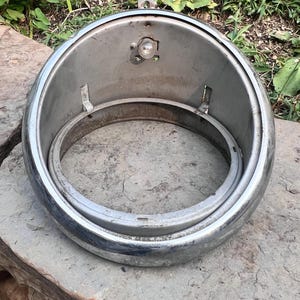 Puede incluir: Una carcasa de faro cromada con un diseño circular. La carcasa tiene una bombilla y soportes de montaje. El objeto está sobre una superficie de piedra.