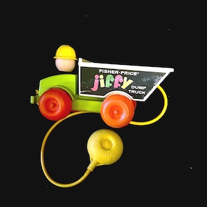 Puede incluir: Un juguete de camión volquete Fisher-Price Jiffy vintage con cuerpo verde lima, ruedas naranjas y una caja negra con el texto blanco Fisher-Price jiffy DUMP TRUCK. Una pequeña figura con casco amarillo está sentada en el asiento del conductor.