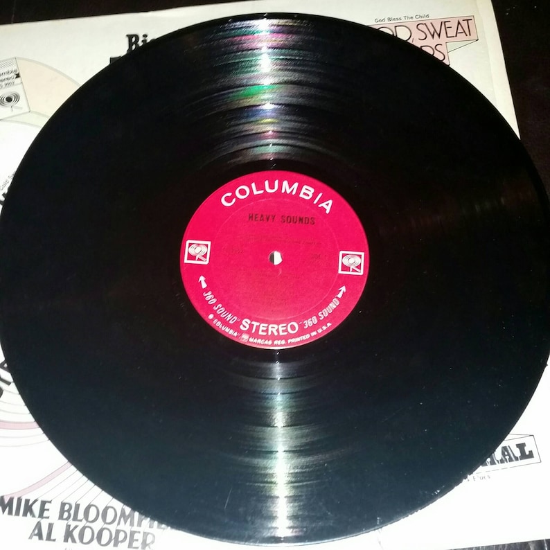 May include: A black vinyl record featuring a circular red center label with white text that reads Columbia, Heavy Sounds, and 360 Sound Stereo. The record is positioned over a white album cover with text including Mike Bloomfield and Al Kooper.
