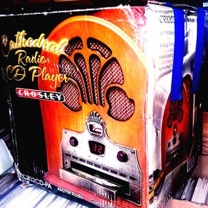 May include: A Crosley Cathedral Radio CD Player box. The box features a vintage radio design with a wooden finish, a silver control panel, and the Crosley logo. The box is secured with blue straps.