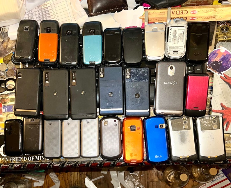 May include: A collection of various used cell phones, including models from LG, Motorola, and Samsung. The phones are in different colors, including black, orange, blue, and silver. Some phones have visible wear and tear.
