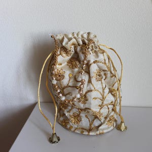 Peut inclure: Une pochette à cordon de couleur crème ornée de broderies florales dorées sur toute la surface. Le sac comprend des cordons dorés terminés par des pompons métalliques en forme de cône et une anse perlée avec de petites perles blanches et dorées.
