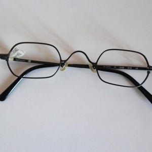 Może przedstawiać: Okulary w czarnej metalowej oprawce z cienkimi, prostokątnymi ramkami i wysokim mostkiem. Okulary posiadają przezroczyste szkła, regulowane noski oraz czarne zauszniki. Na lewym szkle widoczna jest mała biała naklejka.
