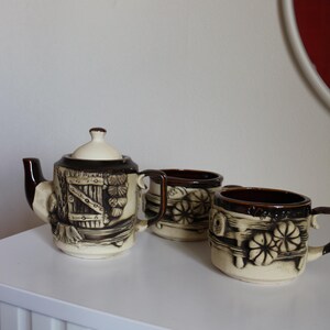 May include: A ceramic tea set consisting of one teapot with a lid and two matching mugs. The pieces are cream-coloured with dark brown accents and feature a raised, embossed relief design depicting a wooden fence, flowers, and foliage.
