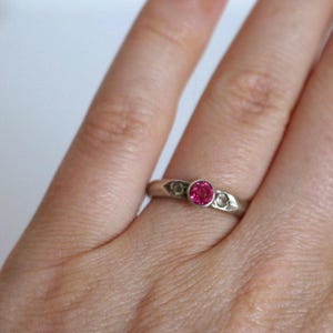 May include: A silver ring with a round, bright pink gemstone in the center, flanked by two smaller, clear stones. The ring is on a finger, showcasing its design and color contrast against the skin.